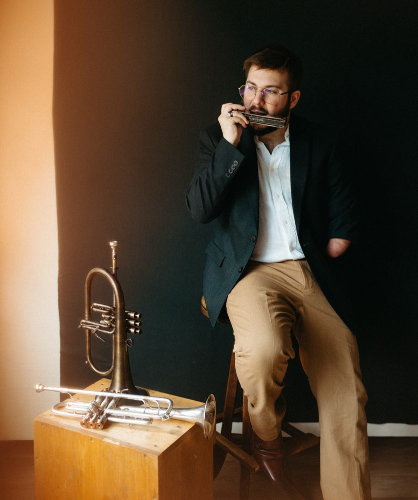 Christian playing chromatic harmonica on a stool, trumpet and flugelhorn on a crate beside him