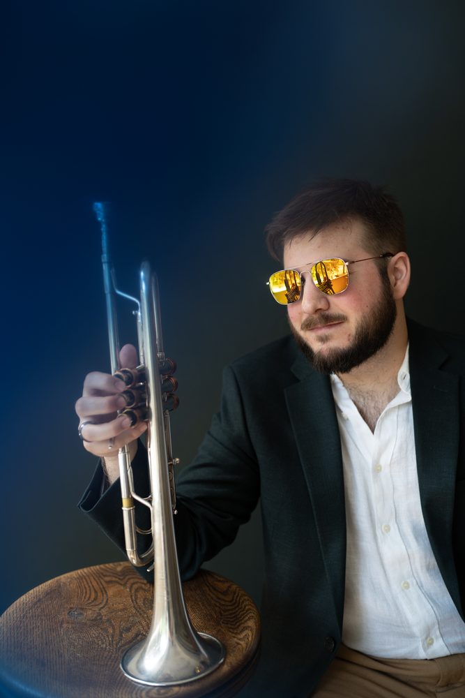 Christian in gold mirrored sunglasses holding a silver trumpet on a wooden stool, deep blue backdrop
